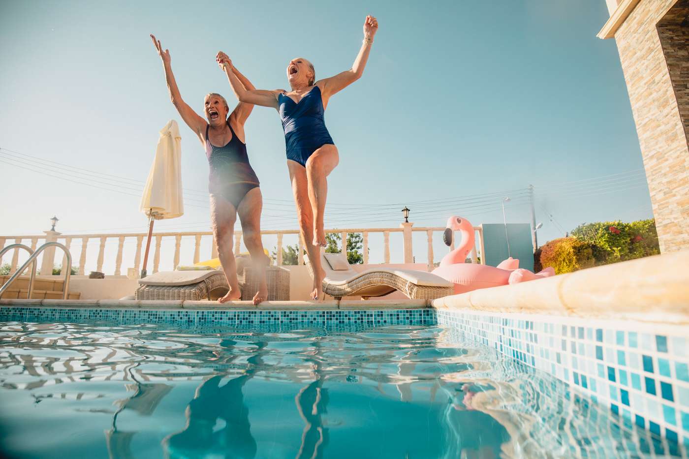 Two senior women holding hands and jumping into a swimming pool. They're excited and having fun.