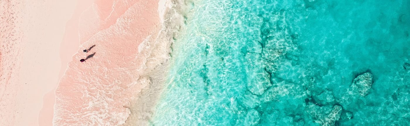 Aerial shot of the beach with two people on the sand and crystal clear seas showing rocks under the surface of the water.